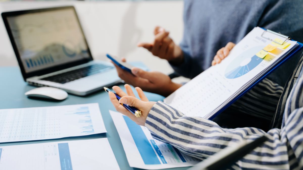 Two people reviewing financial charts and documents at a desk, with a laptop displaying graphs in the background.