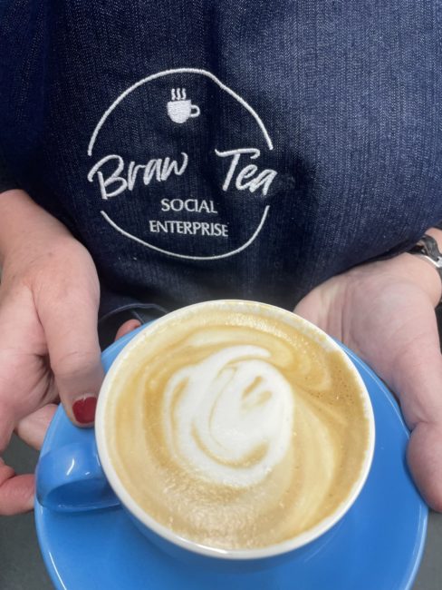Person holding a cup and saucer wearing a Braw Tea branded denim apron
