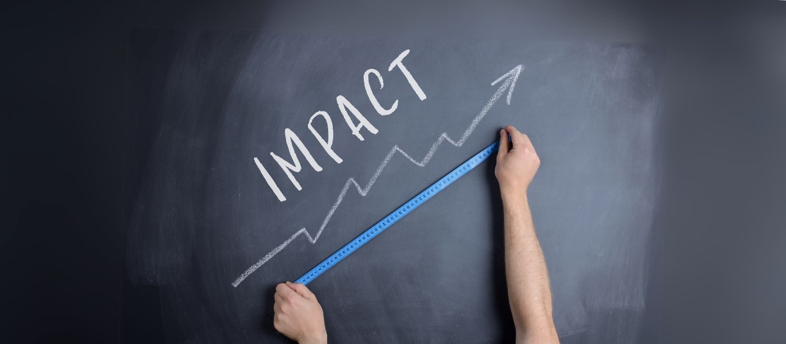A blackboard with the word 'impact' and a diagonal arrow pointing up in chalk. There are hands holding a measuring tape up to the arrow.
