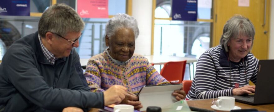 Three older adults sit together at a table using digital devices, smiling and engaged in a relaxed, social setting.