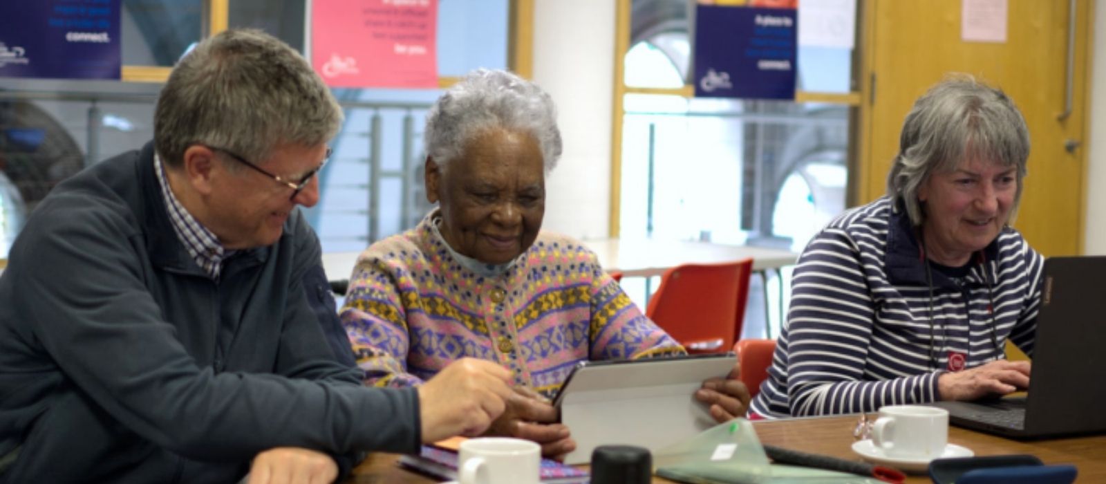 Three older adults sit together at a table using digital devices, smiling and engaged in a relaxed, social setting.