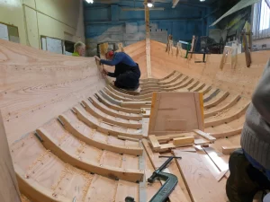 Two people working on the interior wooden frame of a traditional wooden boat under construction.
