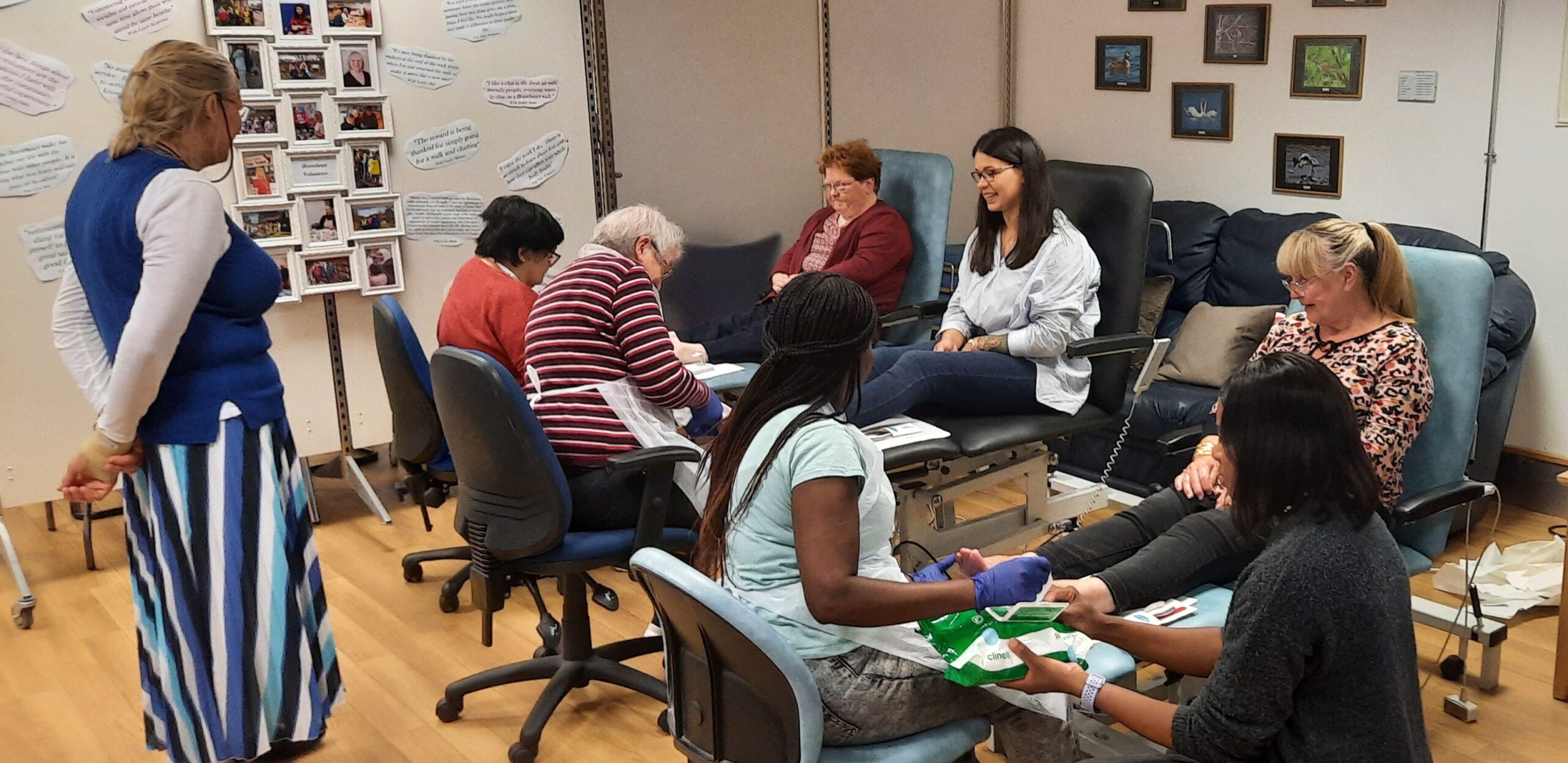 Volunteers provide toenail care to seated clients in a welcoming community setting. The room features comfortable chairs, wall displays, and a relaxed atmosphere.