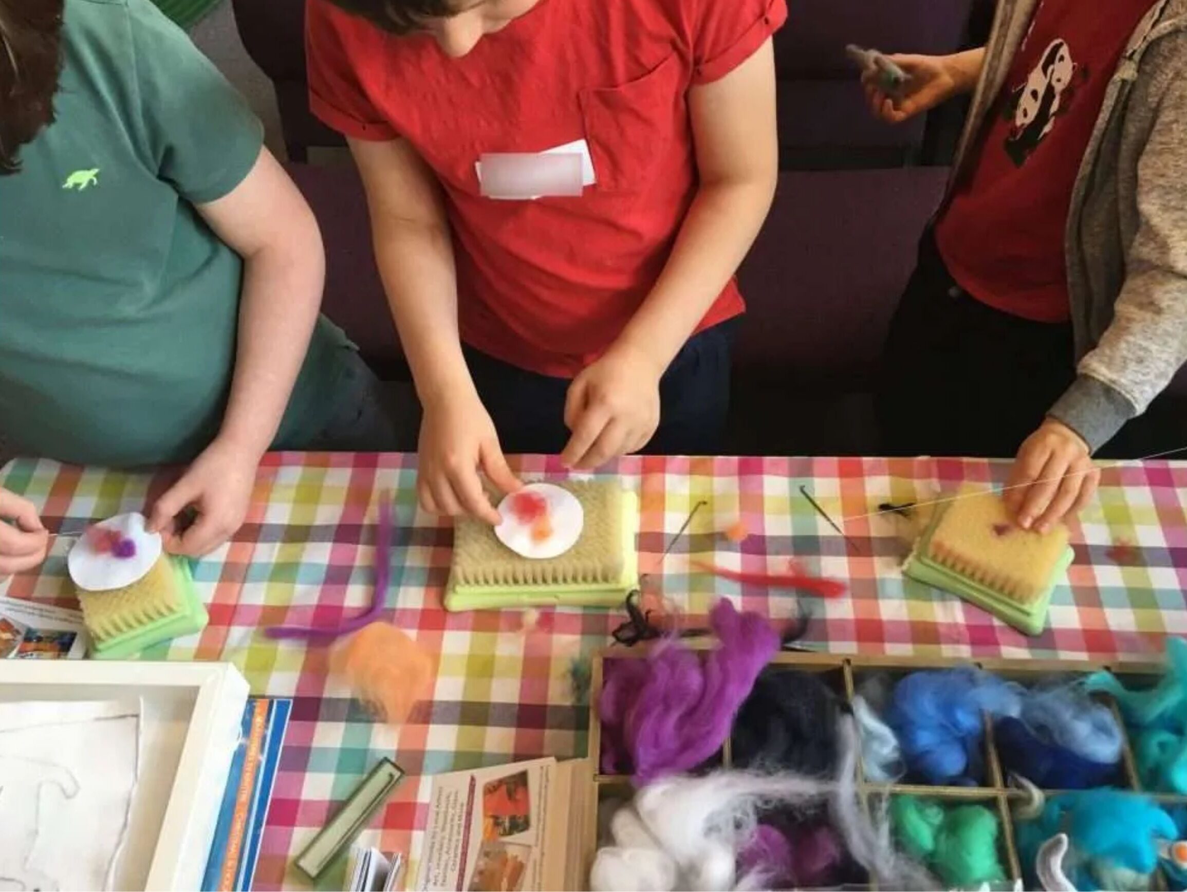 Children crafting with colourful wool fibres at a table covered in a bright checkered cloth.