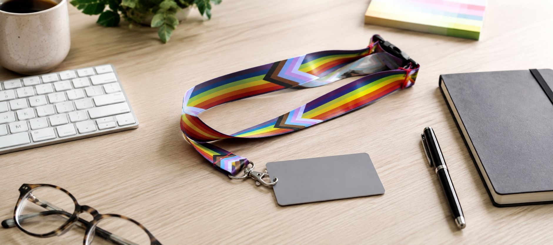 Rainbow Progress Pride lanyard with blank ID badge on a wooden office desk, beside a keyboard, notebook, pen, glasses and coffee cup.